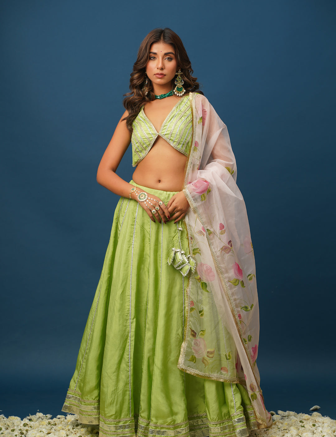 A woman wearing a traditional green lehenga set with a floral design and a dupatta, against a blue background.