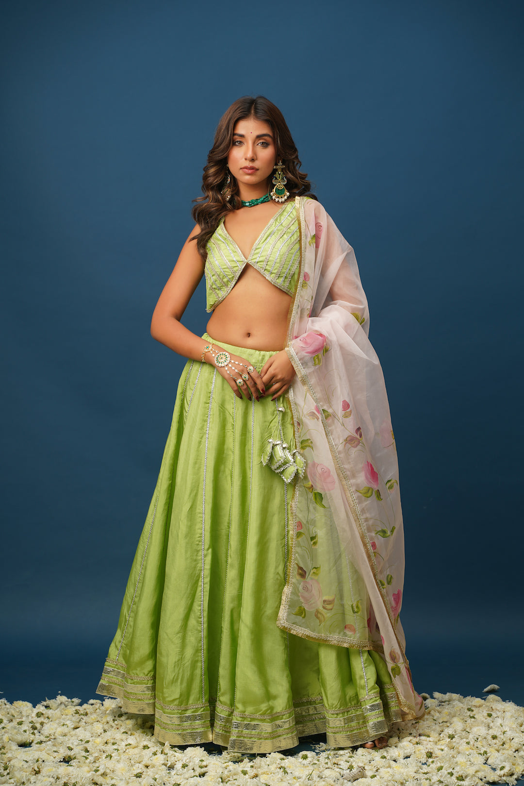 A woman wearing a traditional green lehenga set with a floral design and a dupatta, against a blue background.