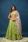 A woman wearing a traditional green lehenga set with a floral design and a dupatta, against a blue background.